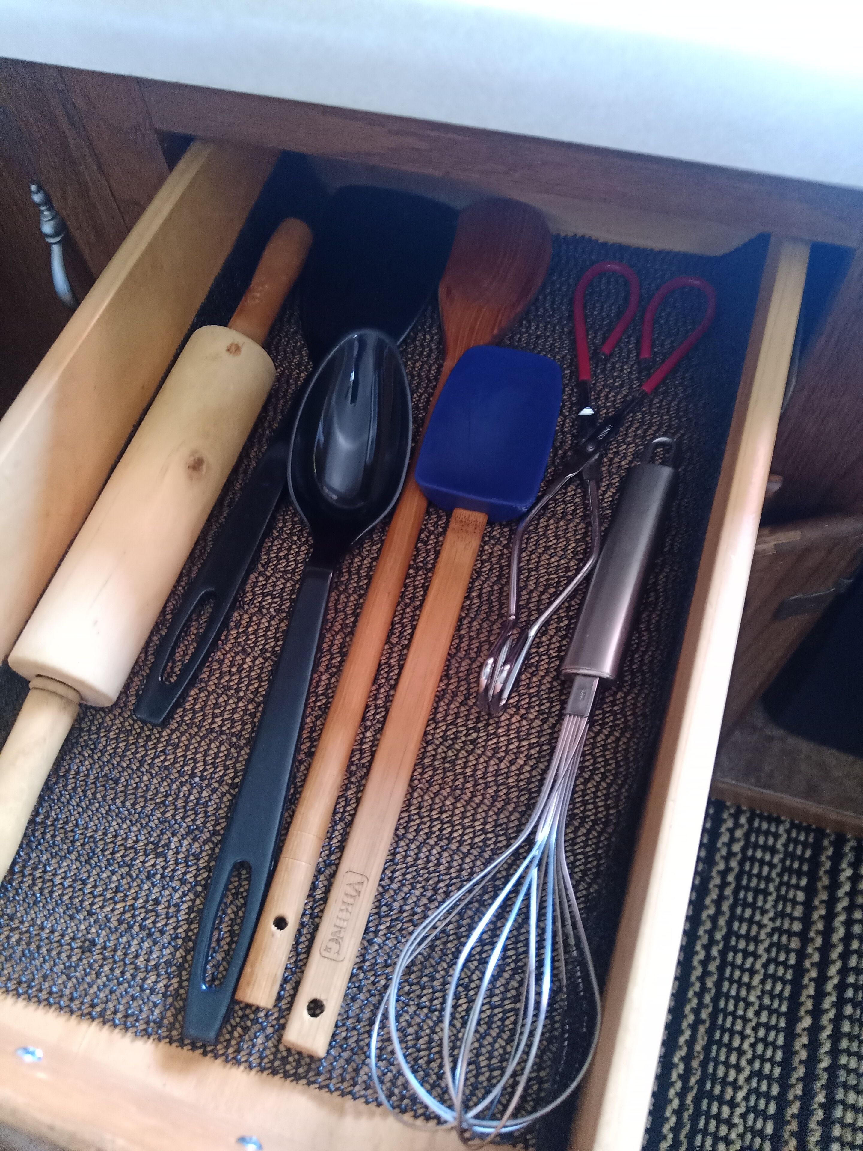A drawer containing kitchen utensils including a rolling pin, a black spoon, a blue spatula, a whisk, and a pair of scissors.
