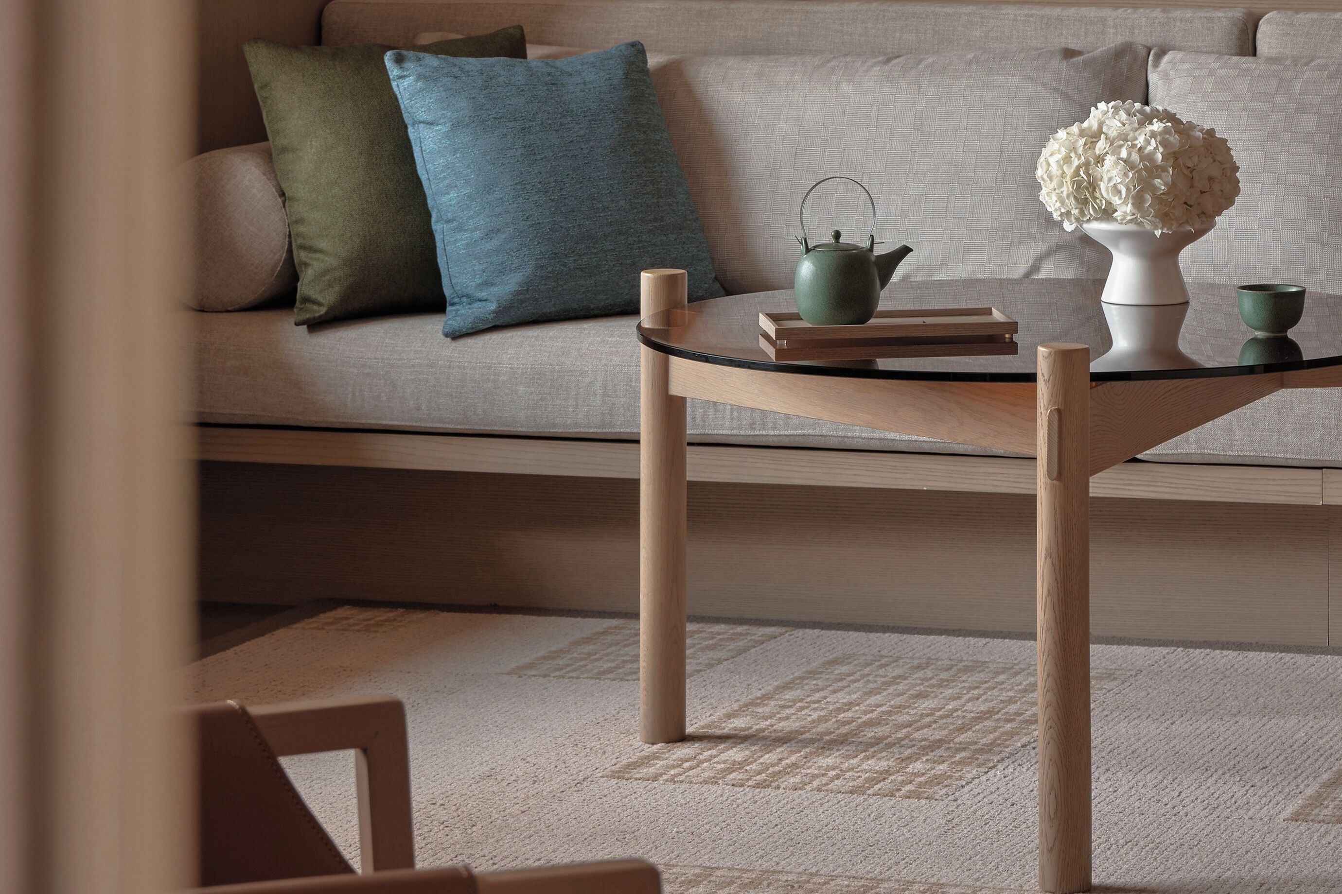 A modern living room with a sofa, pillows, a glass coffee table, a teapot, a vase with flowers, and a cup.