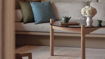 A modern living room with a sofa, pillows, a glass coffee table, a teapot, a vase with flowers, and a cup.