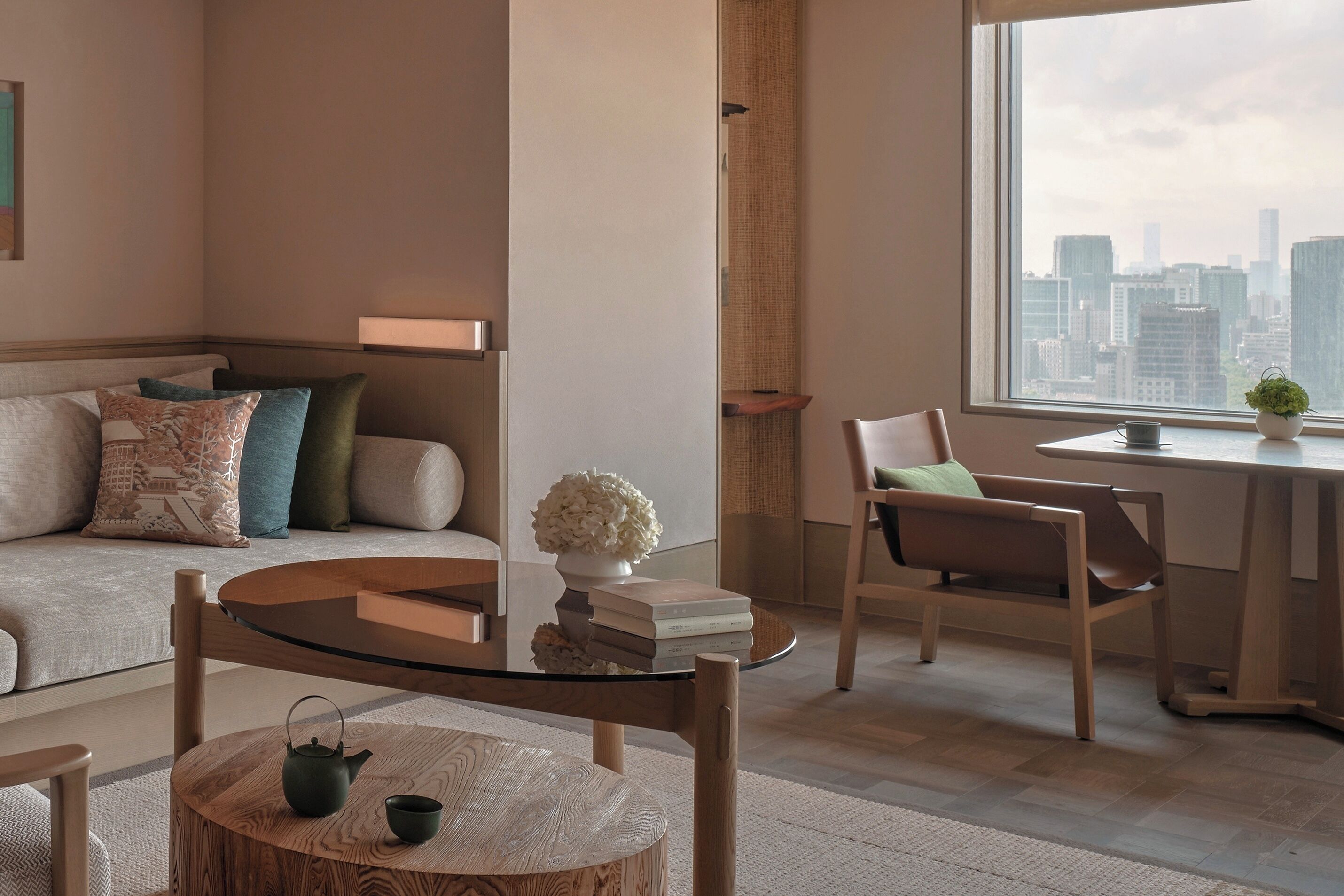 A modern living room with a glass coffee table, a sofa with patterned cushions, a wooden chair, and a view of the city skyline through a large window.