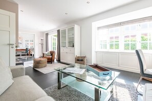 Ein modernes Wohnzimmer mit Glastisch, Ledersessel und Blick auf einen Garten durch große Fenster.