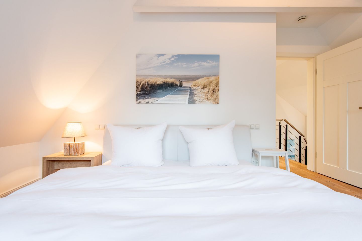 A bedroom with a large bed, a nightstand with a lamp, a painting of a beach scene, and a staircase leading to another room.