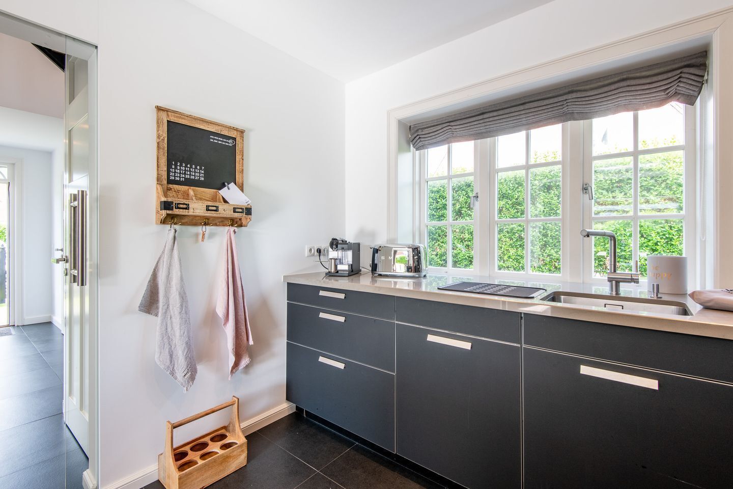 A modern kitchen with dark cabinets, a built-in oven, a coffee machine, and a window with a view of greenery.