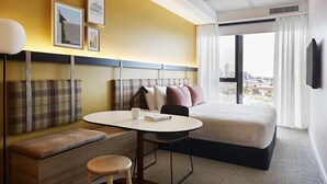 Habitación de hotel con cama, una pequeña mesa con silla, un banco y vistas de la ciudad a través de amplios ventanales.