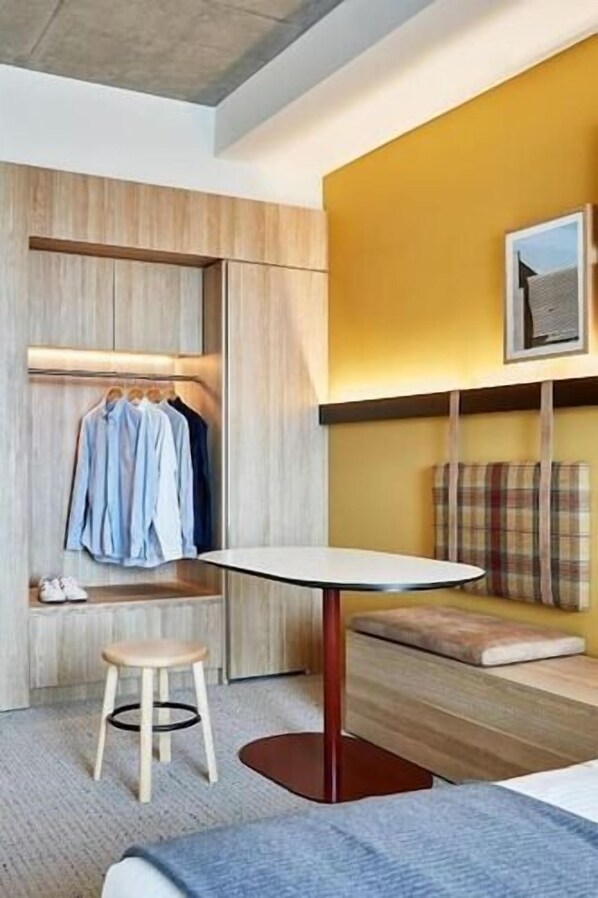 A modern room with a wooden wardrobe, a small stool, a round table, and a yellow wall with a framed picture.