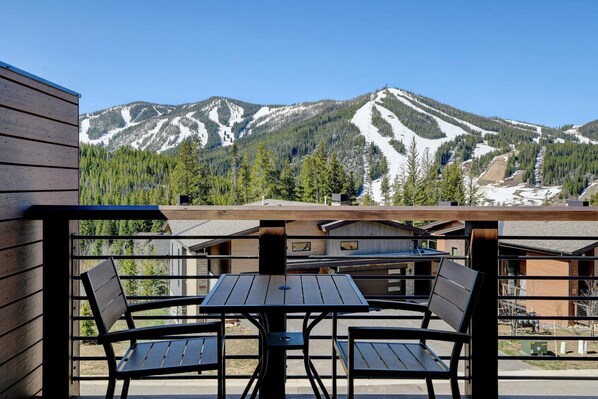 A wooden deck with a table and chairs, overlooking a mountain with ski slopes and forested areas.