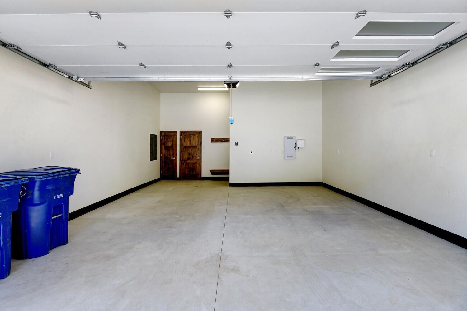 En hall med ett betonggolv, vita väggar och en trädörr längst ner. På vänster sida finns blå soptunnor och på höger vägg sitter en ljusströmbrytare.