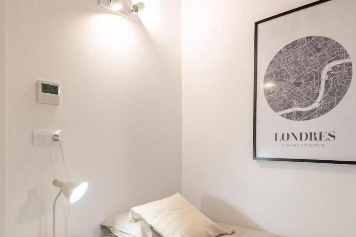 A bedroom with a bed, a nightstand with a lamp, a wall-mounted light, and a framed cityscape picture.