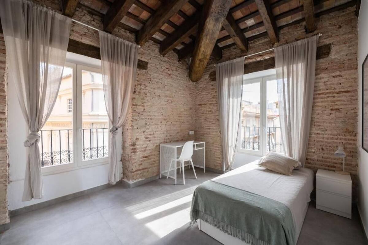 A bedroom with exposed brick walls, a wooden ceiling, a bed, a desk, and a window with curtains.