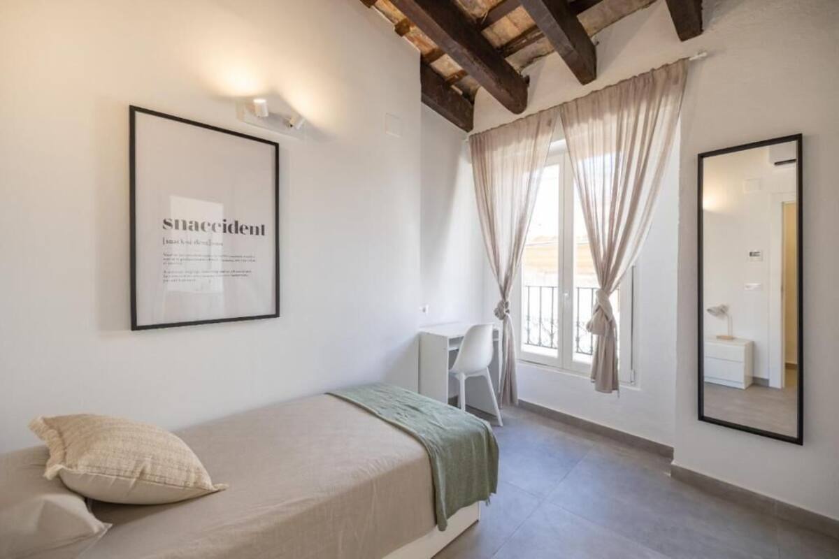 A small, well-lit bedroom with a single bed, a desk, and a large mirror.