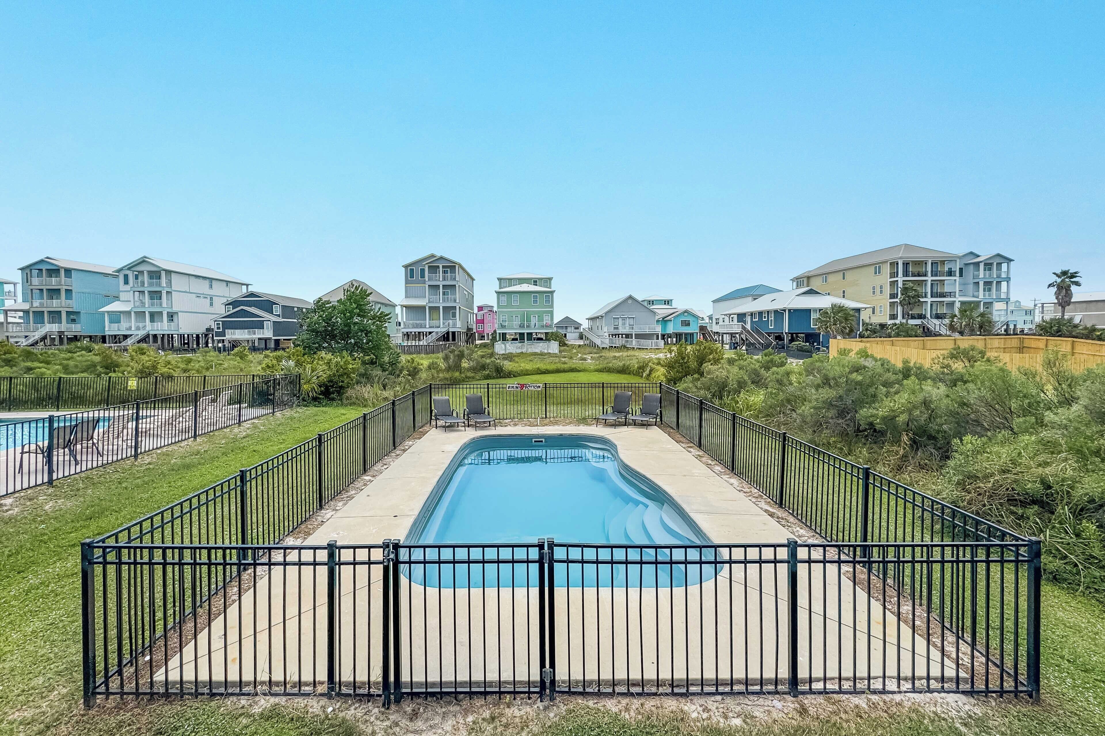 A swimming pool surrounded by a black metal fence, with a grassy area and a row of colorful buildings in the background.