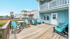 Une terrasse en bois avec des fauteuils Adirondack turquoise et une petite table, d’où l’on peut admirer une plage et d’autres bâtiments.