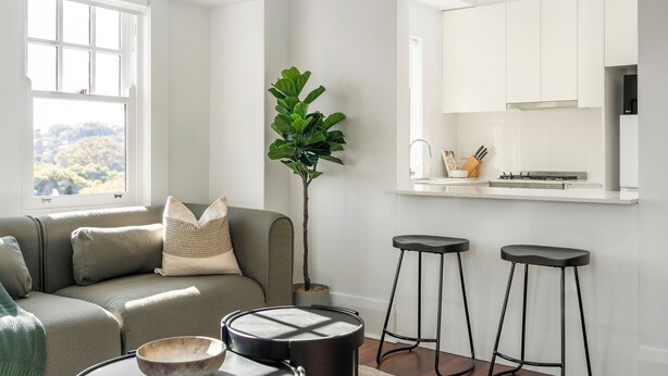 Una sala moderna con un sofá, una mesa de centro y una planta. Hay dos taburetes y una zona de cocina con armarios y un lavamanos.