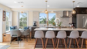 A modern kitchen with a marble countertop, a central island with bar stools, and large windows offering a view of the outdoors. - Slow M'Ocean – A Fresh Take on Cape Calm (Chatham)