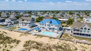 A coastal residential area with houses, a large blue-roofed building with a pool, and a beach in the foreground. - Over the Moon | Oceanfront | Private Pool and Hot Tub (Virginia Beach)