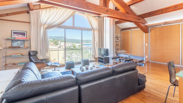 A spacious living area with a large sofa, a coffee table, and a television. There is a balcony with a view of the landscape.