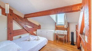A bedroom with a sloped ceiling, wooden beams, a bed with white linens, a desk with a chair, and a radiator.