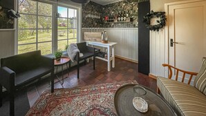 Een gezellige kamer met een groot raam, een kleine tafel met een glazen karaf, een stoel, een bank en een kamerkleed met een patroon.