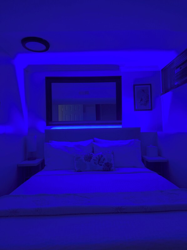 Un dormitorio con cama, una mesita de noche, un televisor montado en la pared y un cuadro enmarco.