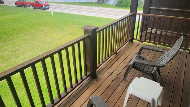 Terraza de madera con dos sillas Adirondack y una mesa pequeña, con vista a un césped y una carretera con vehículos.