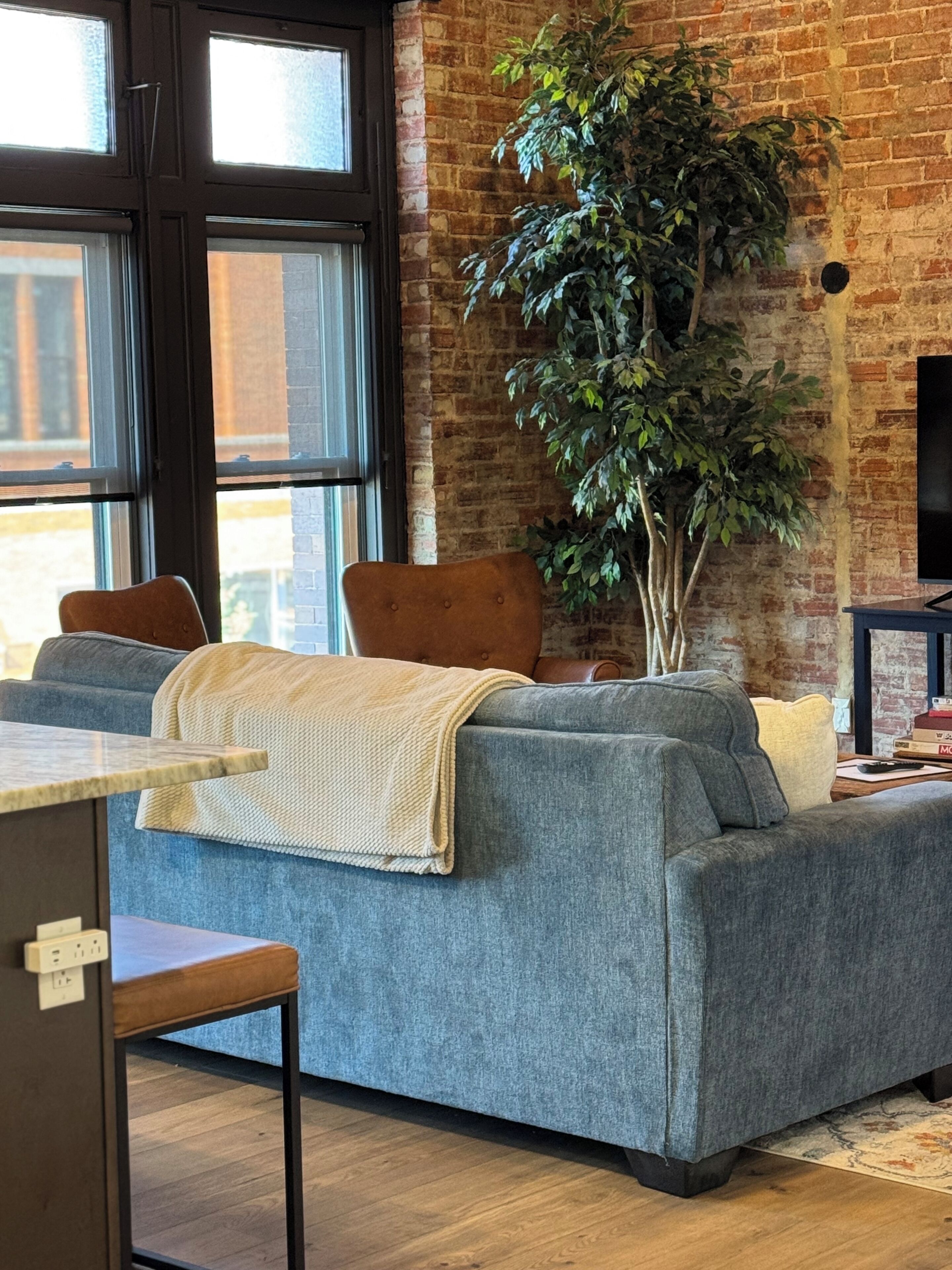 A living room with a grey sofa, a large potted plant, and a television against a brick wall.