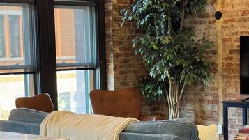 A living room with a grey sofa, a large potted plant, and a television against a brick wall.