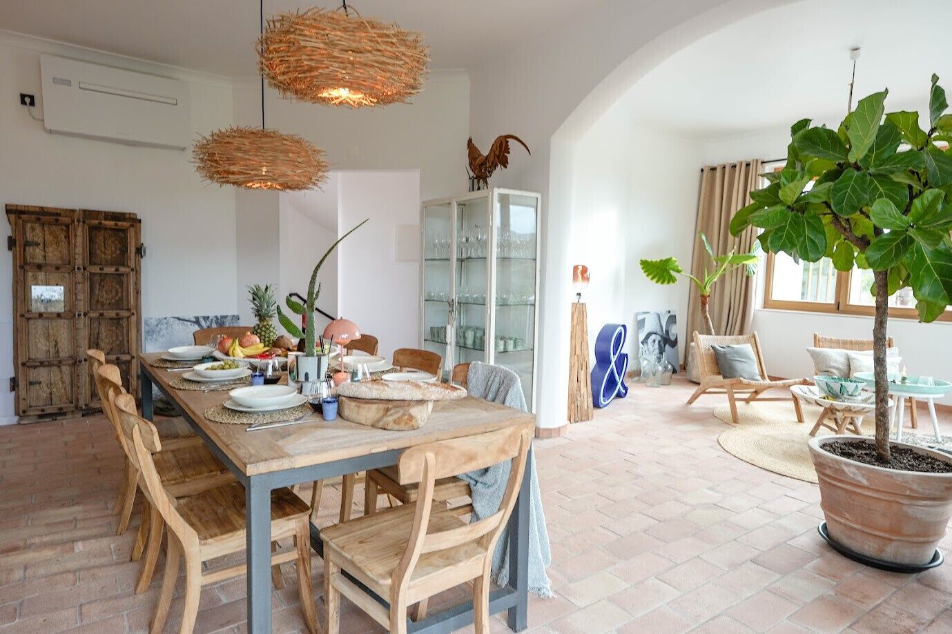 Ein geräumiger Essbereich mit einem Holztisch, der für eine Mahlzeit gedeckt ist, umgeben von Holzstühlen. In der Ecke steht ein großer Blumentopf und eine Vitrine mit Dekorationen.