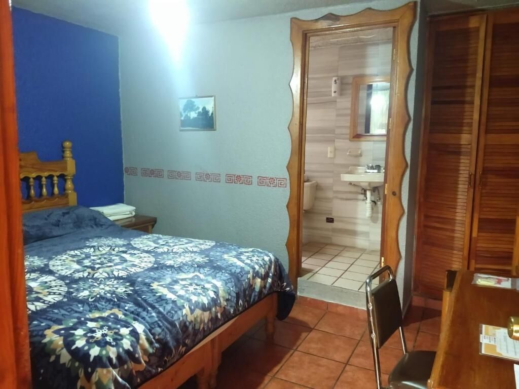 Un dormitorio con una cama, cabecera de madera, pared azul, una puerta que da al baño y una pequeña mesa con una silla.