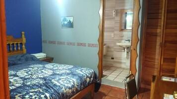 Un dormitorio con una cama, cabecera de madera, pared azul, una puerta que da al baño y una pequeña mesa con una silla.