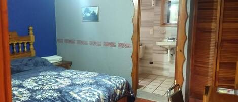 Un dormitorio con una cama, cabecera de madera, pared azul, una puerta que da al baño y una pequeña mesa con una silla.