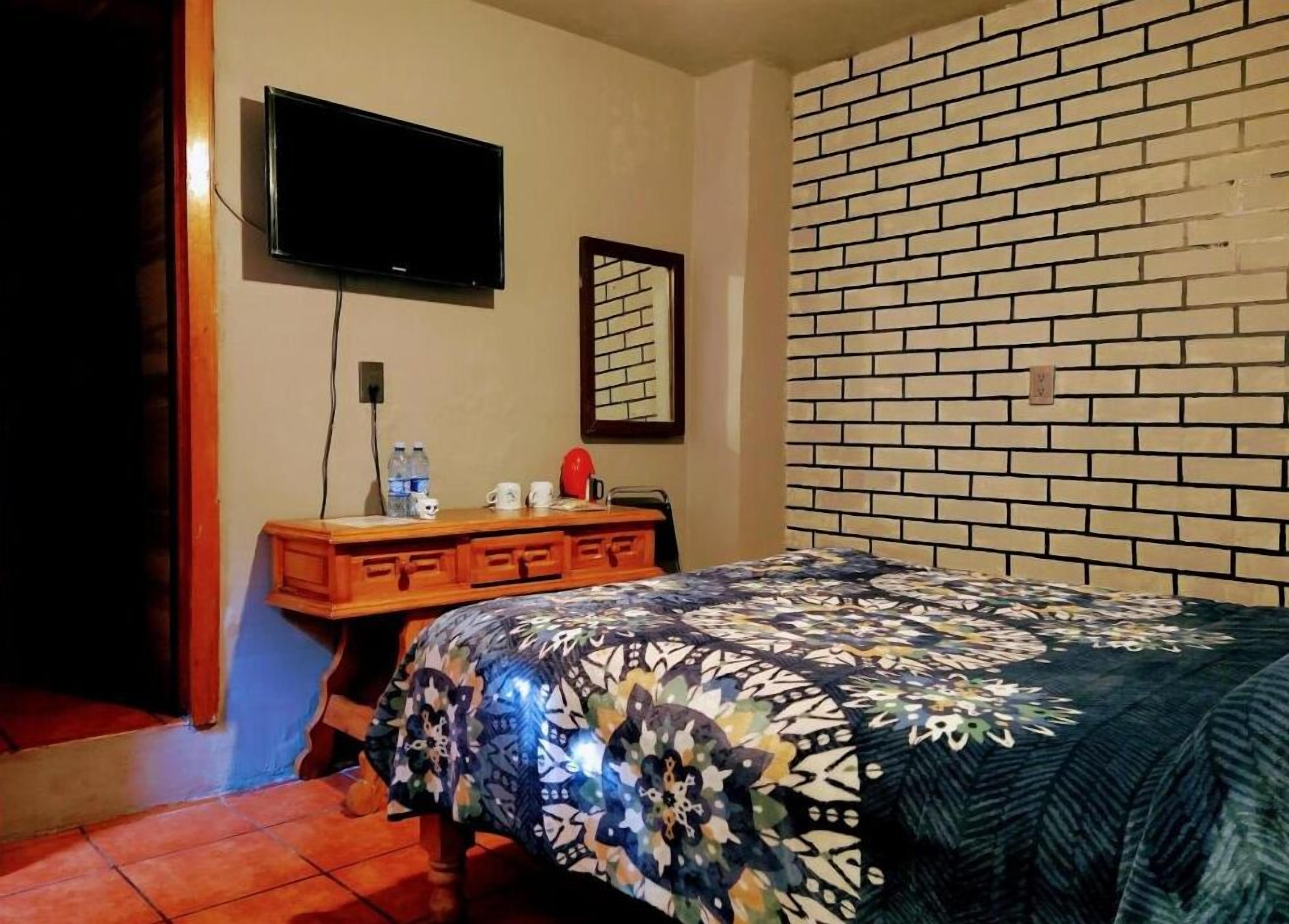 Een slaapkamer met een wand in baksteenmotief, een bed met een bedovertrek met dessin, een houten nachtkastje met spiegel, en een televisie die aan de muur is bevestigd.