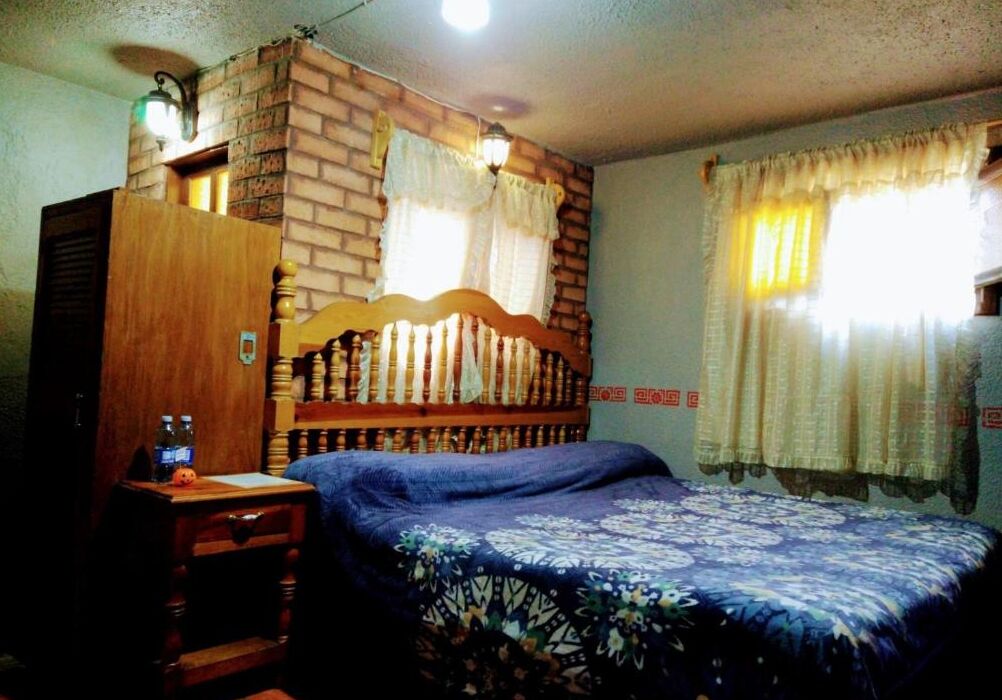Un dormitorio con cabecera de madera, cama con colcha estampada, mesita de noche con una botella y pared de ladrillo.