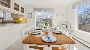 A dining room with a wooden table set for four, white chairs, a vase with flowers, and a view of trees through the window.