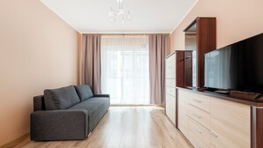 Een moderne woonkamer met een grijze bank, houten vloeren, een tv op een houten kast en een groot raam met gordijnen.