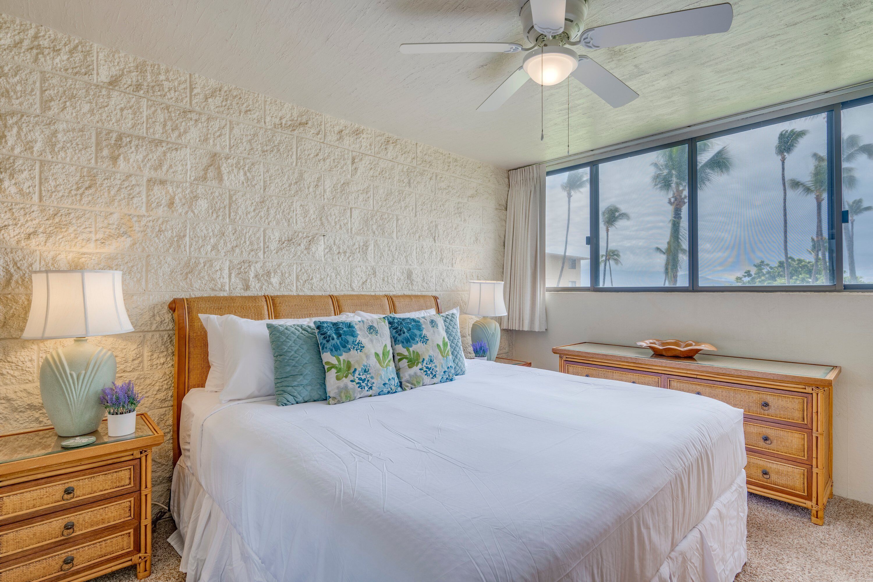 Quarto com cama grande, cabeceira de madeira, duas mesas de cabeceira com luminárias, ventilador de teto e vista para palmeiras pela janela.