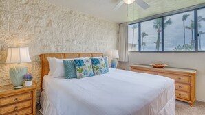 Ein Schlafzimmer mit einem großen Bett, einem Holzkopfende, zwei Nachttischen mit Lampen, einem Deckenventilator und Blick auf Palmen durch ein Fenster.