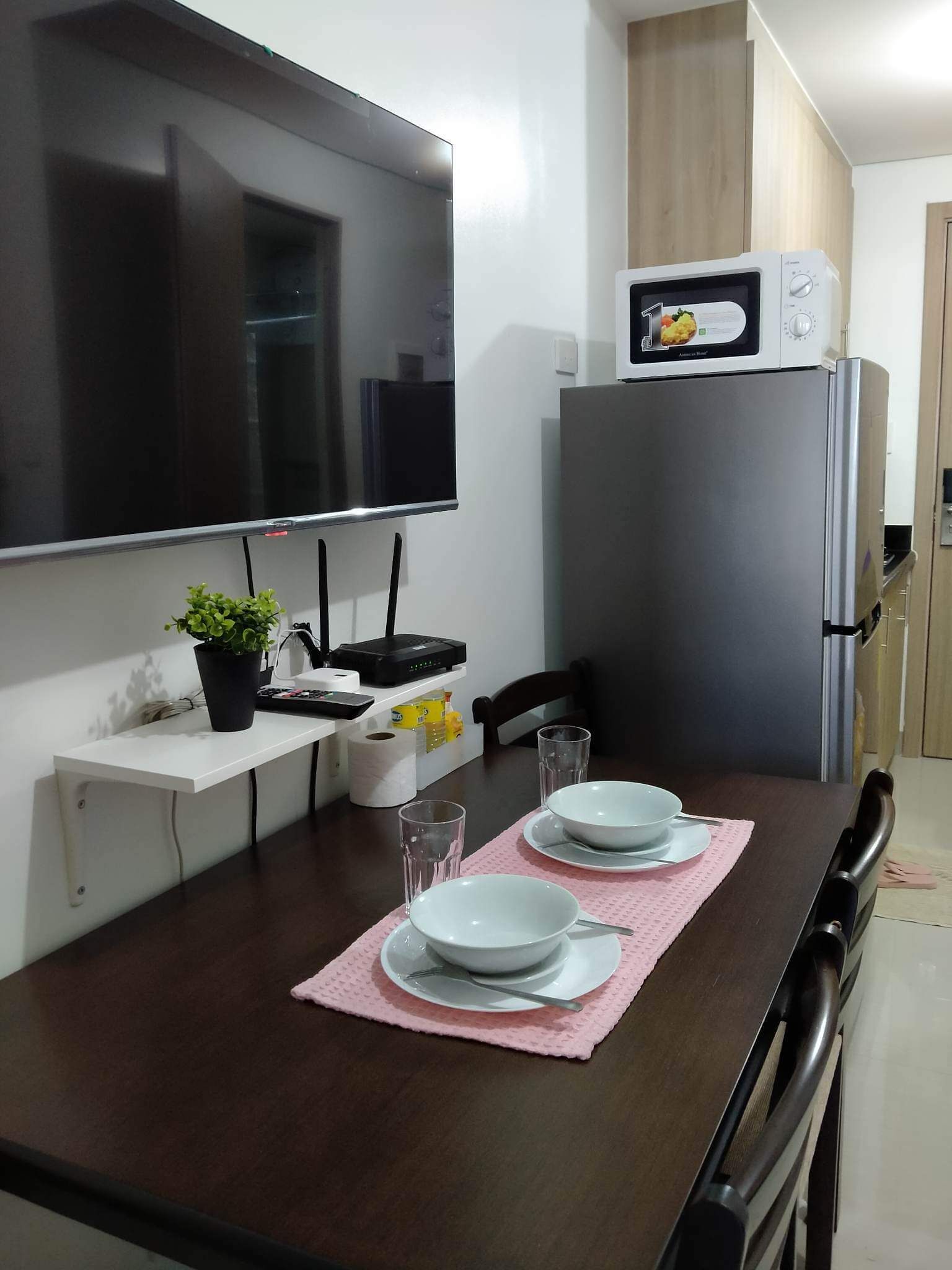 A modern kitchen with a flat-screen TV, a microwave, and a refrigerator. There is a dining table with plates and glasses, and a small plant on a shelf.