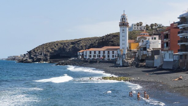Un pueblo costero con una torre blanca destacada, costa rocosa y edificios con balcones.