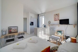 A modern hotel room with a bed, a desk with a chair, a TV, a small kitchen area, and a window.