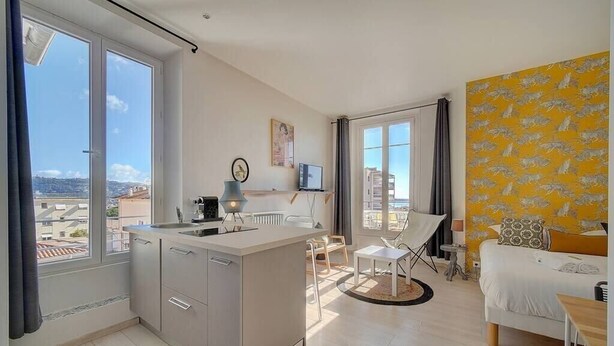 A modern apartment with a kitchen, dining area, and a bedroom. It features large windows, a view of the city, and a yellow patterned wall. - Upstays Studio Top Floor Sea View Cannes (Cannes)