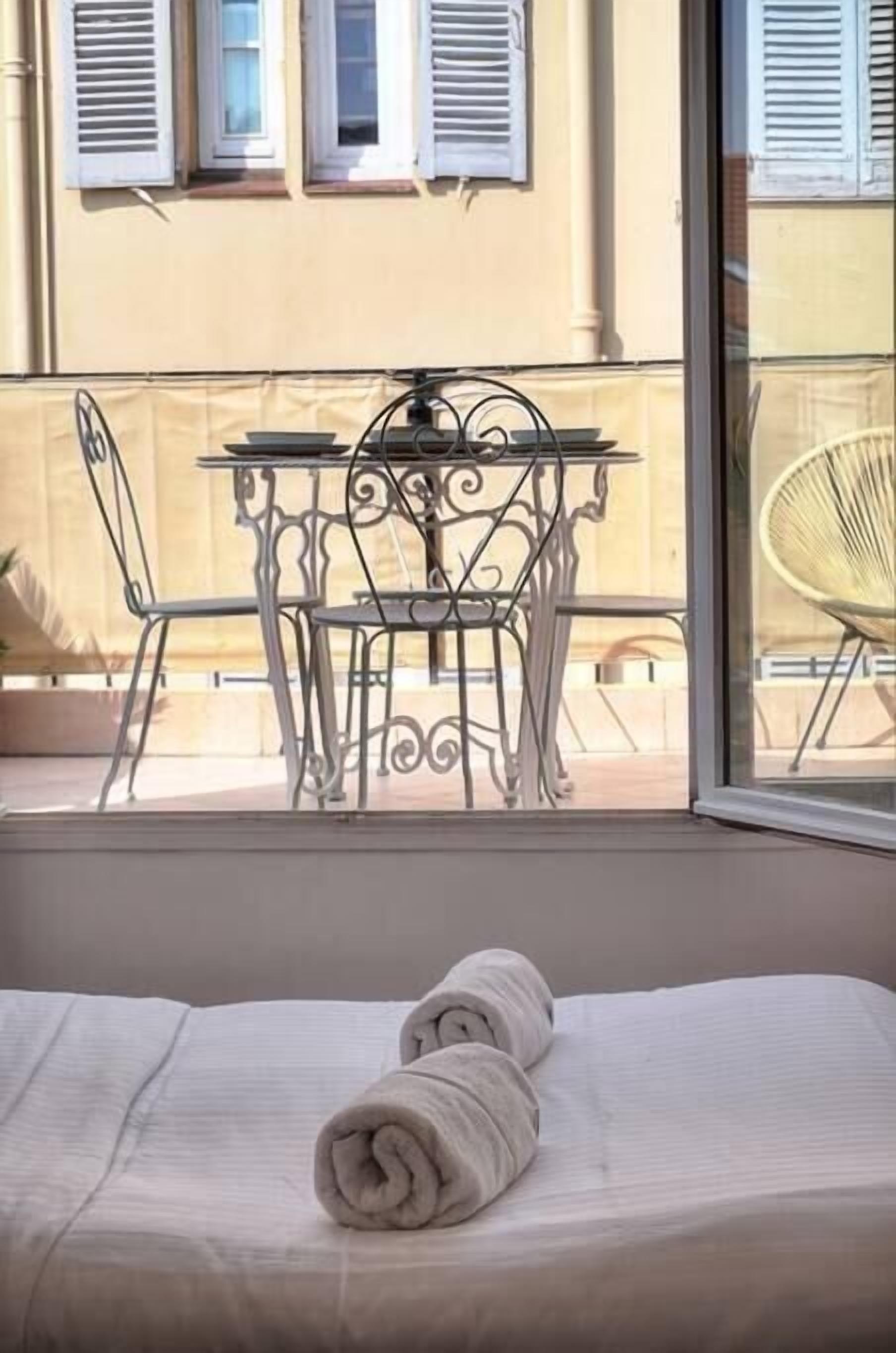 A balcony with a table and chairs, a bed with a rolled towel, and a building with shutters in the background.