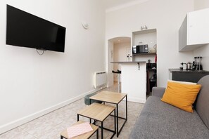 Una sala de estar moderna con un televisor de pantalla plana, un sofá con un cojín mostaza, una máquina de café y una cocina con microondas.