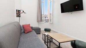 Een moderne woonkamer met een grijze bank, een kleine tafel, een groene poef, een vloerlamp en een aan de muur bevestigde televisie.