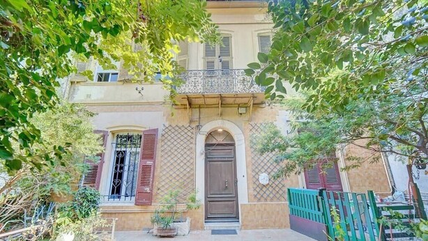 Un edificio de dos pisos con un balcón, una puerta grande de madera y un pequeño jardín.