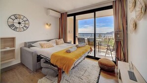 A modern bedroom with a bed, a bedside table, a small sofa, a large window with a view, and a wall clock. - Upstays Terrace Pool Parking 40m² Apartment (Cannes)