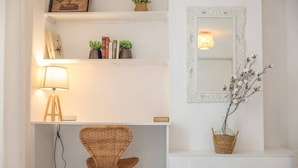A room with a white wall, a wicker chair, a white table, a lamp, a mirror, and shelves with books and plants. - Upstays Palais Center 38m² Studio (Cannes)