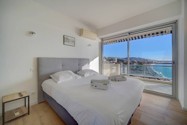 Un dormitorio con una cama grande, una mesita de noche, aire acondicionado instalado en la pared y vista al mar a través de una puerta corrediza de vidrio.