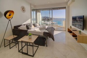 Una sala de estar moderna con sofá, una mesa de centro y televisor. Hay un balcón con vista al mar.