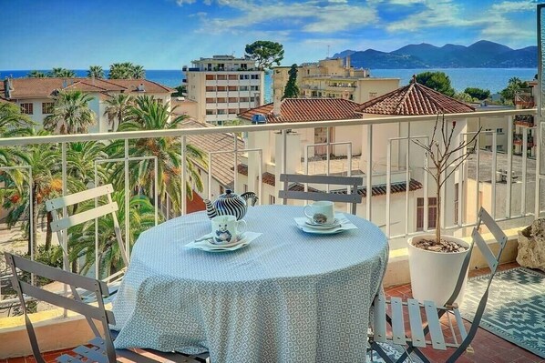A balcony with a table set for two, overlooking a coastal cityscape with buildings and a view of the sea.