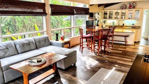 A modern living room with a grey sofa, wooden coffee table, and dining area with wooden chairs. Large windows offer a view of greenery outside. - Roof deck views, hot tub, big kitchen, 3200sf, BBQ (Kailua-Kona)